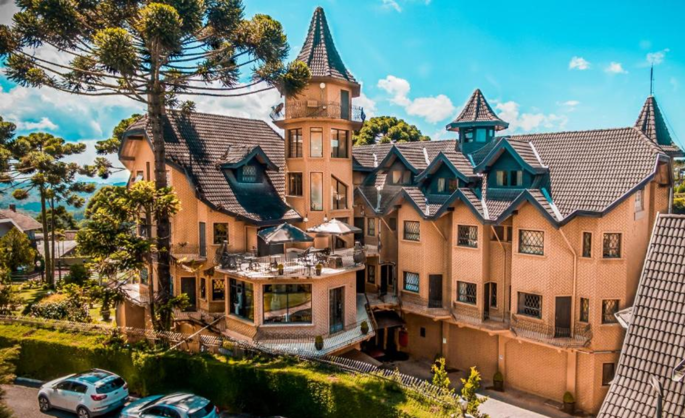 Fachada do Hotel Castelo Nacional Inn em Campos do Jordão ao entardecer com torres iluminadas, névoa nas montanhas ao fundo e jardins com flores da serra em primeiro plano